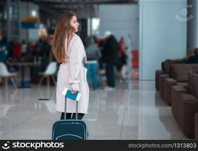 Young female traveller walking the airport hall draging suitcase. Blured airport cafe on the background. Suitable for bus, railway, metro station.