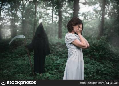 Young female victim in white dress in the forest, death in a black hoodie and with a scythe on background. Photo in horror style, exorcism. Young female victim in white dress in the forest
