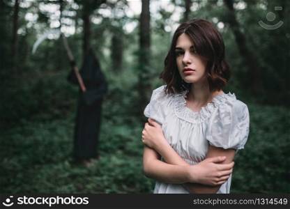 Young female victim in white dress in the forest, death in a black hoodie and with a scythe on background. Photo in horror style, exorcism. Young female victim in white dress in the forest