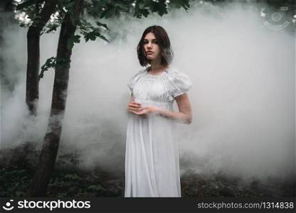 Young female victim in white dress in the misty forest. Photo in horror style, mystery. Female victim in white dress in the misty forest