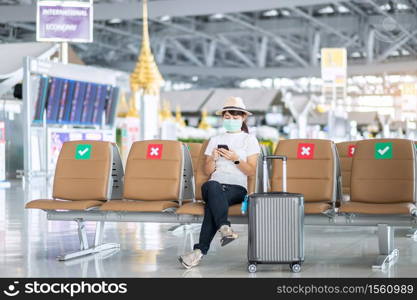 Young female wearing face mask and using mobile smartphone in airport, protection Coronavirus disease (Covid-19) infection, Asian woman traveler sitting on chair. New Normal and social distancing