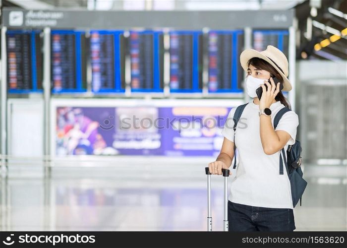 Young female wearing face mask and using mobile smartphone in airport terminal, protection Coronavirus disease infection, Asian woman traveler with hat. New Normal and travel under COVID-19 concept