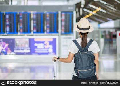 Young female wearing face mask with luggage checking flight time in airport, protection Coronavirus disease infection, Asian woman traveler ready to travel. New Normal and travel under COVID-19