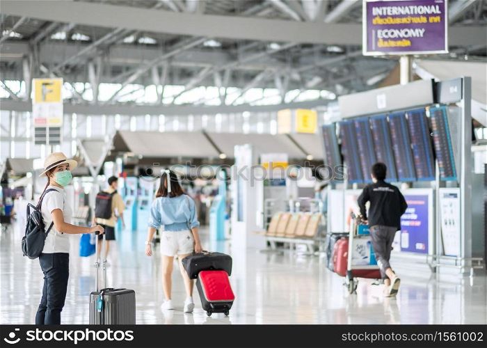 Young female wearing face mask with luggage walking in airport terminal, protection Coronavirus disease (Covid-19) infection, Asian woman traveler with hat. New Normal and travel bubble concept