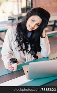 Young female with dark hair holding takeaway coffee sitting in front of opened laptop browsing internet. Glad woman with long hair dressed formally working with computer. Technology and youth concept