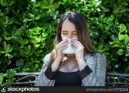 Young female with hay fever sneezing with closed eyes and wiping nose with paper handkerchief while resting near green shrub in park. Woman with pollen allergy sneezing with closed eyes