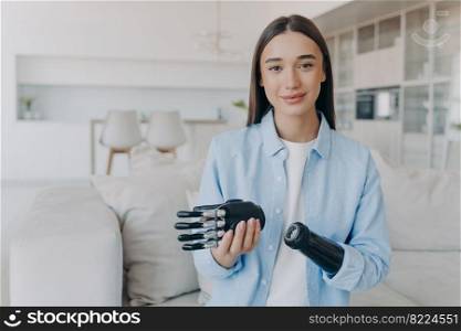 Young female with prosthetic arm showing her high tech bionic prosthesis at home. Girl installing artificial limb, looking at camera. Modern people with disabilities, new medical technologies concept.. Young girl with prosthetic arm showing high tech bionic prosthesis. Modern people with disabilities