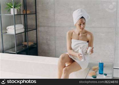Young female wrapped in towel applying moisturizing skincare cosmetics after taking a shower, sitting in bathroom. Woman taking care of body skin after bathing. Self-care, spa procedure.. Female applying moisturizing skincare cosmetics for body skin after taking a shower in bathroom
