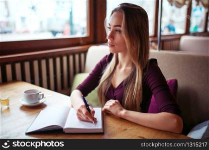 Young female writing in notebook against window in restaurant.