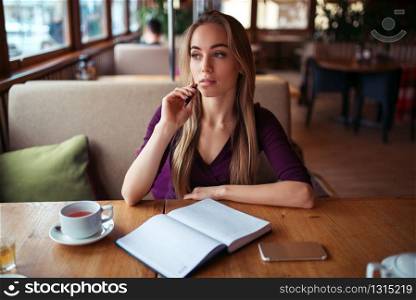Young female writing in notebook against window in restaurant.. Female writing in notebook in restaurant.