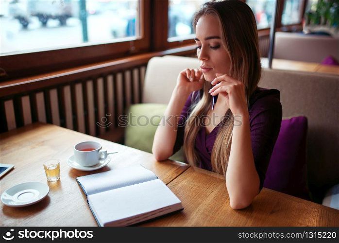 Young female writing in notebook against window in restaurant.. Female writing in notebook in restaurant.
