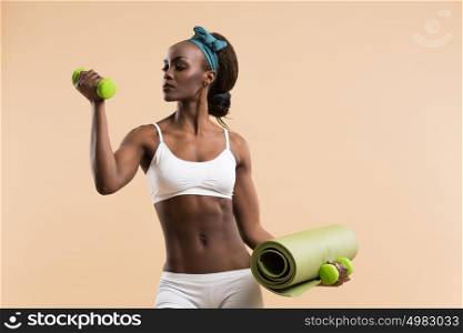 Young fit african woman with dumbbells and yoga mat on beige background