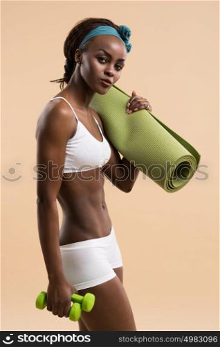 Young fit african woman with dumbbells and yoga mat on beige background