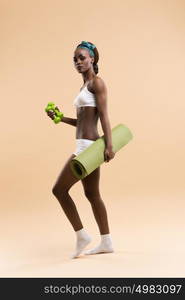 Young fit african woman with dumbbells and yoga mat on beige background