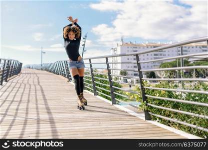Young fit black woman on roller skates riding outdoors on urban bridge with open arms. Smiling girl with afro hairstyle rollerblading on sunny day. Beautiful clouds in the sky.