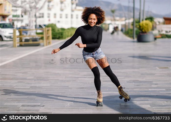 Young fit black woman on roller skates riding outdoors on urban street. Smiling girl with afro hairstyle rollerblading on sunny day