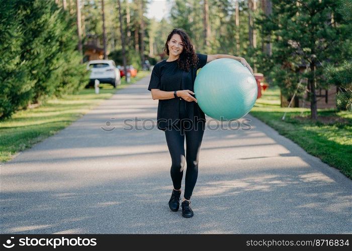 Young fit brunette woman holds big fitness ball, dressed in active wear, does exercises workout, poses in park, walks outdoor and breathes fresh air. People, gymnastics and aerobics concept.