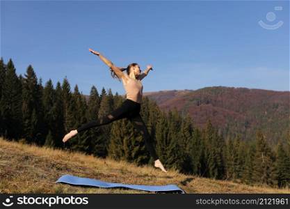 Young fit woman doing exercises on mat on background of sunny mountains hills. Outdoor workout, healthy lifestyle. female in sport suit is jumping in nature against the forest in the morning.. Young fit woman doing exercises on mat on background of sunny mountains hills. Outdoor workout, healthy lifestyle. female in sport suit is jumping in nature against the forest in the morning
