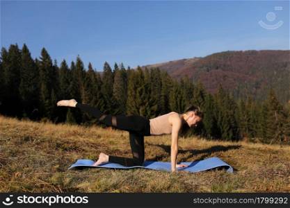 Young fit woman doing exercises on yoga mat on background of sunny mountains hills. Outdoor workout, healthy lifestyle. female in sport suit doing asanas in nature against the forest in the morning.. Young fit woman doing exercises on yoga mat on background of sunny mountains hills. Outdoor workout, healthy lifestyle. female in sport suit doing asanas in nature against the forest in the morning