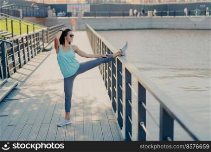 Young fit woman with dark hair stretches legs on fence warms up before jogging wears sunglasses t shirt leggings and sneakers prepares for cardio training poses outdoor being in good physical shape