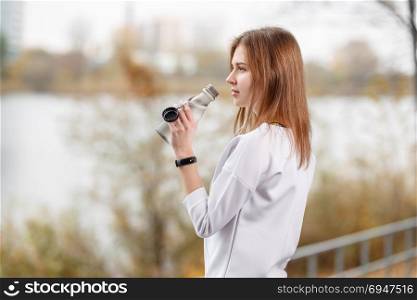 Young fitness girl with bottle of water standing outdoor in city park.. Young fitness girl with bottle of water standing outdoor in city park
