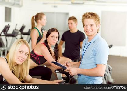 Young fitness instructor with gym people spinning or at cross-trainer