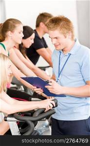 Young fitness instructor with gym people spinning or at cross-trainer