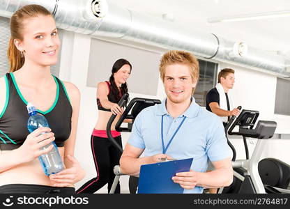 Young fitness instructor with gym people spinning or at cross-trainer