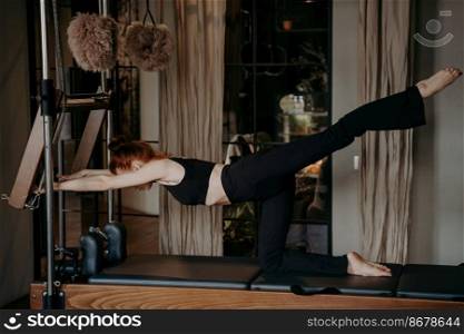 Young flexible woman exercising and stretching leg back on reformer during pilates training in cozy fitness studio with relaxing atmosphere, wearing sportswear. Female doing stretching exercises. Young athletic woman exercising and stretching leg on pilates reformer