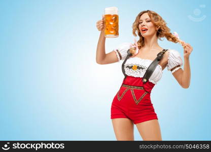 Young flirting sexy Swiss woman wearing red jumper shorts with suspenders in a form of a traditional dirndl, holding a beer mug on blue background.