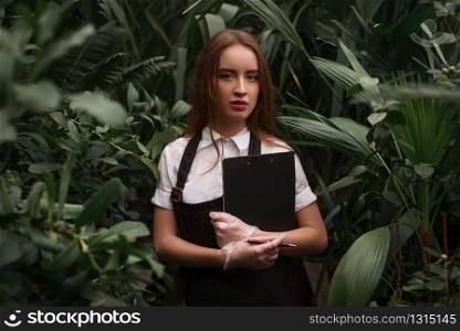 Young florist in apron holds clipboard in hands against greenhouse with exotic plants.. Florist in apron holds clipboard in hands