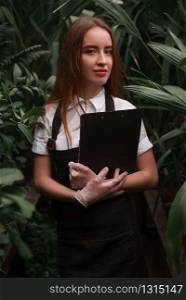 Young florist in apron holds clipboard in hands against greenhouse with exotic plants.