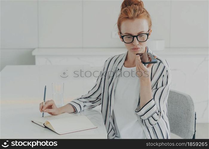 Young focused concentrated ginger woman freelancer using voice recognition system on smartphone, making notes in notebook, recording audio message while sitting at table in kitchen, working remotely. Young focused concentrated ginger woman freelancer using voice recognition system on smartphone