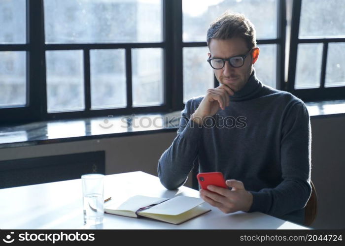 Young focused german businessman wearing glasses using mobile phone while sitting at desk with opened notebook, handsome austrian man holding smartphone, reading internet news or checking email. Young german businessman reading internet news or checking email on smartphone while sitting at desk