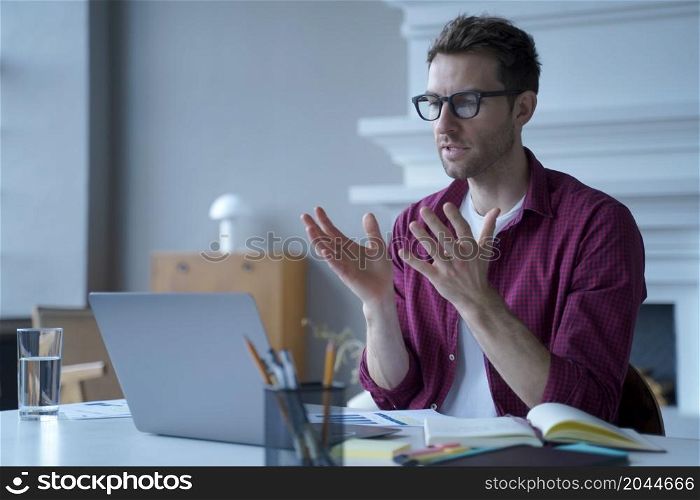 Young focused german male entrepreneur in glasses and casual clothes participating in virtual online meeting with colleagues, making video call on laptop computer while working in modern office. Young focused german man in glasses and casual clothes participating in virtual online meeting