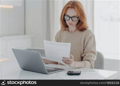 Young focused red-haired woman in spectacles calculating budget at home, sitting at table with laptop, holding paper taxes bills and reading financial information, female managing family budget. Young focused red-haired woman sitting at table with laptop and holding paper taxes bills