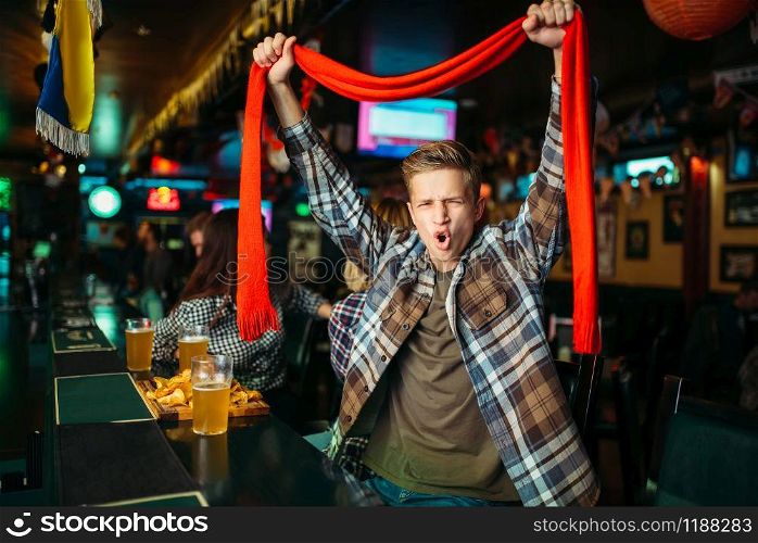 Young football fan with scarf raises his hands up at the counter in sports bar. Tv broadcasting, teenager watching the match of the favorite team, success game celebration in pub