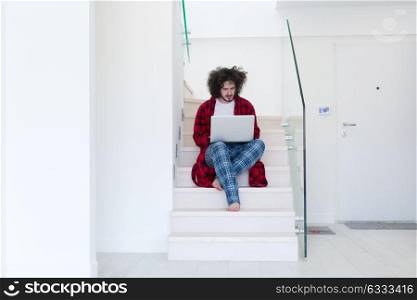 young freelancer in bathrobe working from home using laptop computer while sitting on stairs