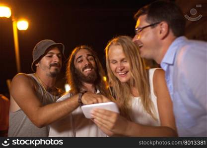 Young friends laughing at what they see on mobile. Group of friends having fun outdoor at night. They looking at smart phone and laughing. Funny photos or humorous video