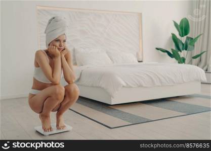 Young frustrated female barefoot in underwear squatting on electronic smart scales during morning weighing after shower procedure to find out her result. Healthy lifestyle and dieting concept. Young sad and unhappy girl in underwear barefoot in underwear squatting on electronic smart scales