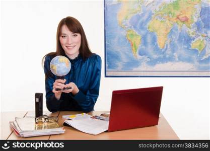 Young geography teacher sitting behind a desk, behind which hangs a map of the world