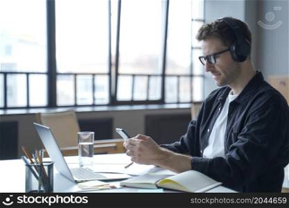 Young german male freelancer in headphones and glasses looking at smartphone display in hands, checking last emails or typing message sms while sitting at workplace in office. Young german male freelancer in headphones and glasses looking at smartphone display in hand