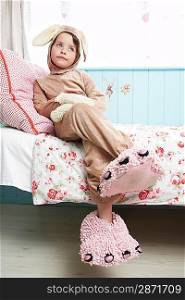 Young girl (5-6) sitting on bed wearing bunny costume and monster slippers
