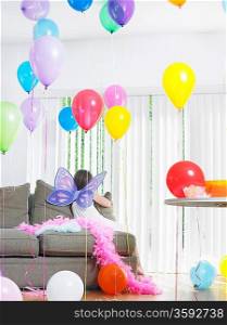 Young girl (7-9) sitting on sofa with balloons wearing fairy wings back view