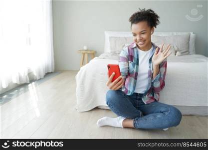 Young girl answer video call on phone wave hand to camera, greeting friend. Smiling biracial female blogger streaming online in social network, welcoming followers, sitting on bedroom floor.. Smiling biracial girl blogger wave hand to phone camera greeting followers in social network at home