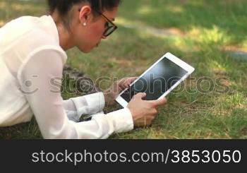 Young girl browsing her tablet laying on the grass in the park.Clerk chatting during her leisure time.Businesswoman sending message with tablet.Leisure typing woman an email on her iPad.