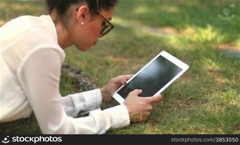 Young girl browsing her tablet laying on the grass in the park.Clerk chatting during her leisure time.Businesswoman sending message with tablet.Leisure typing woman an email on her iPad.