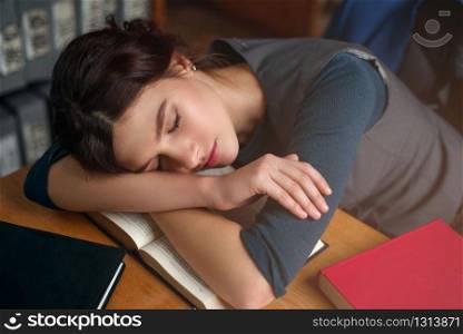 Young girl fell asleep while reading a book in college library. Bookshelf on the background. Student examination concept.