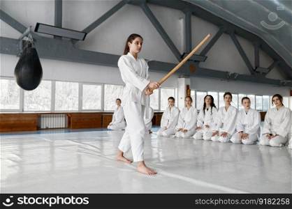 Young girl fighting with wooden sword at aikido training in martial arts school. Teenage female fighter in white kimono showing technique to her classmates. Young girl fighting with wooden sword at aikido training in martial arts school