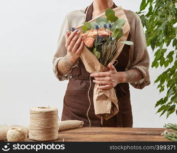 Young girl florist with tattoo hands hold bouquet with fresh flowers roses living coral color in a paper on a gray wall background. Concept floral shop and small business. Mother's Day holiday.. Woman florist with fresh beautiful handmade bouquet from fesh fragrant flowers ang green leaves on a light wall background, copy space.